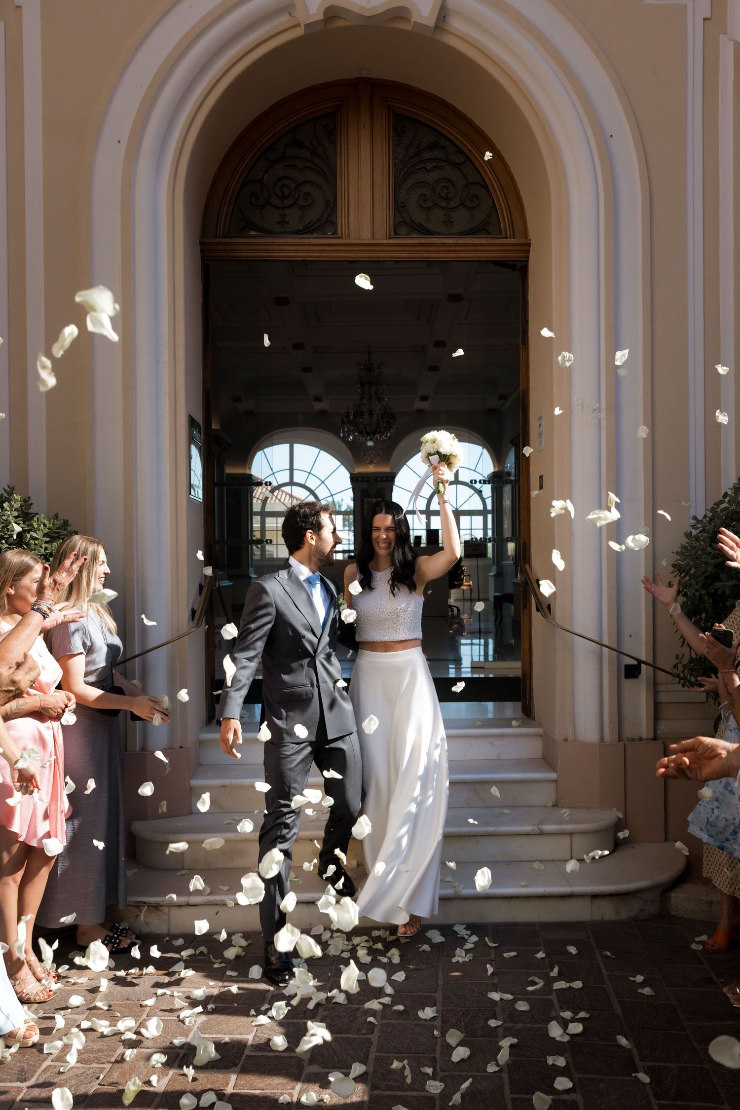 Bride and groom exit church amid white flower petal confetti shower, guests celebrating their wedding ceremony.