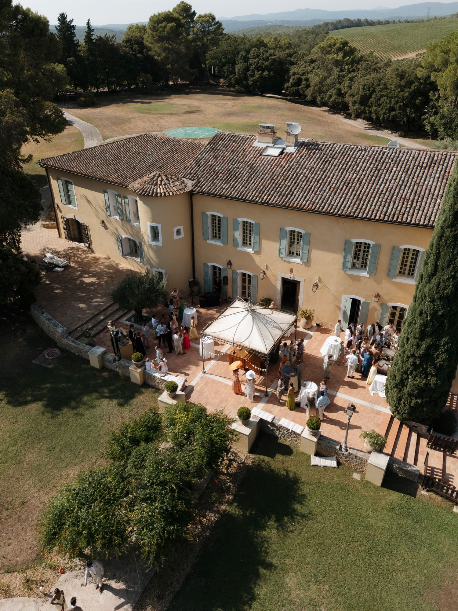 Aerial view of a elegant Provençal villa with terracotta roof hosting an outdoor wedding reception surrounded by lush greenery.