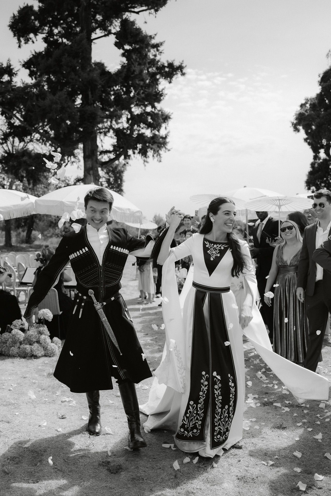 Black and white photo of a bride and groom in traditional Georgian attire walking joyfully at an outdoor wedding ceremony.