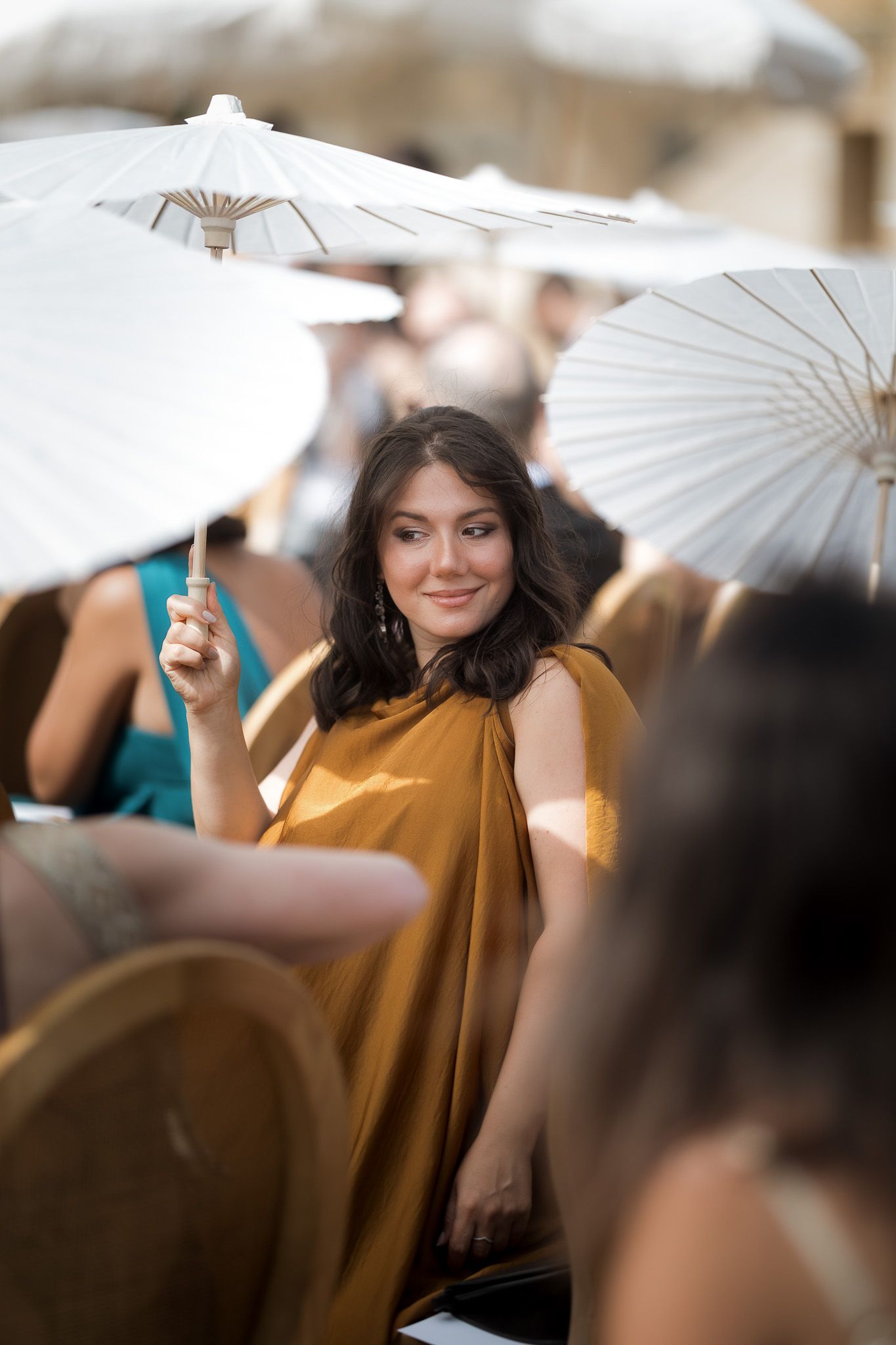 A woman in a golden dress holds a white parasol umbrella at an outdoor event.