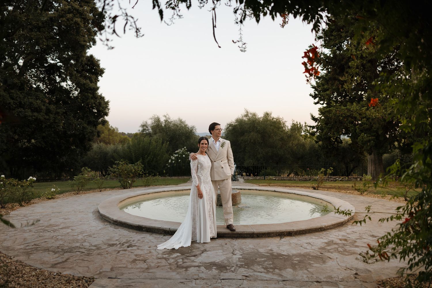 Bride and groom standing by a stone fountain in a lush garden at dusk, bride in white gown, groom in tan suit.