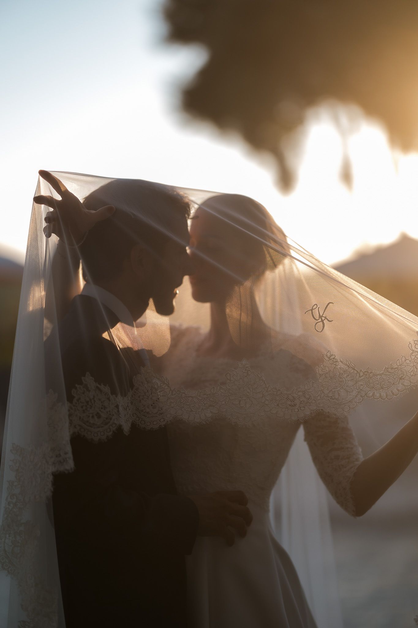 Bride and groom sharing intimate moment under wedding veil at golden sunset, romantic silhouette portrait.