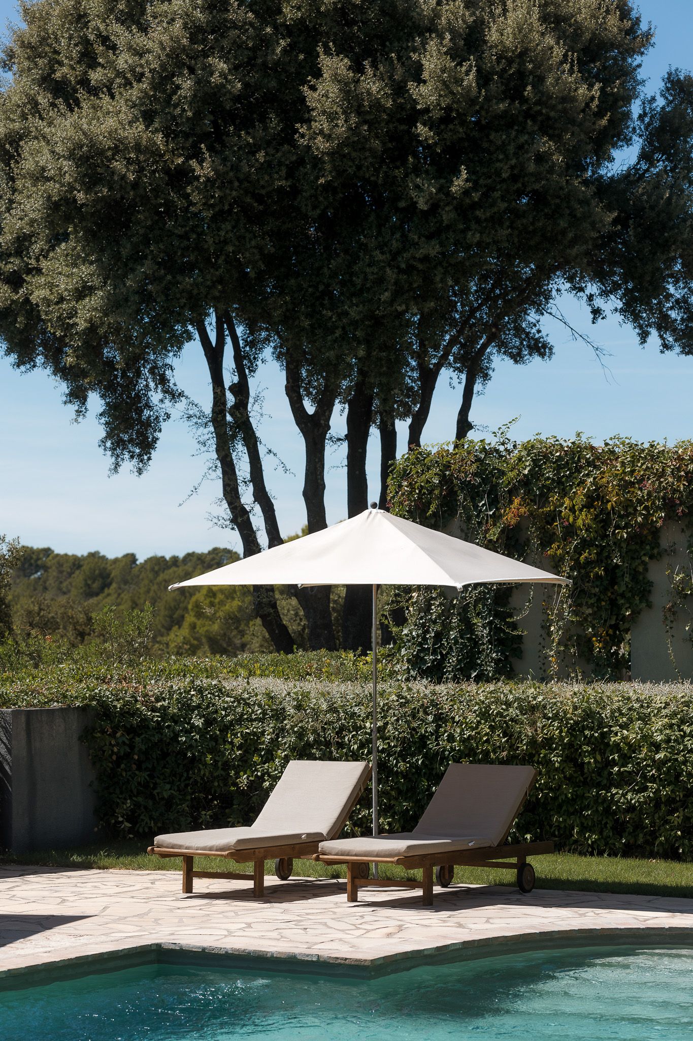 Two wooden lounge chairs under a white umbrella beside a pool, surrounded by lush hedges and trees on a sunny day.