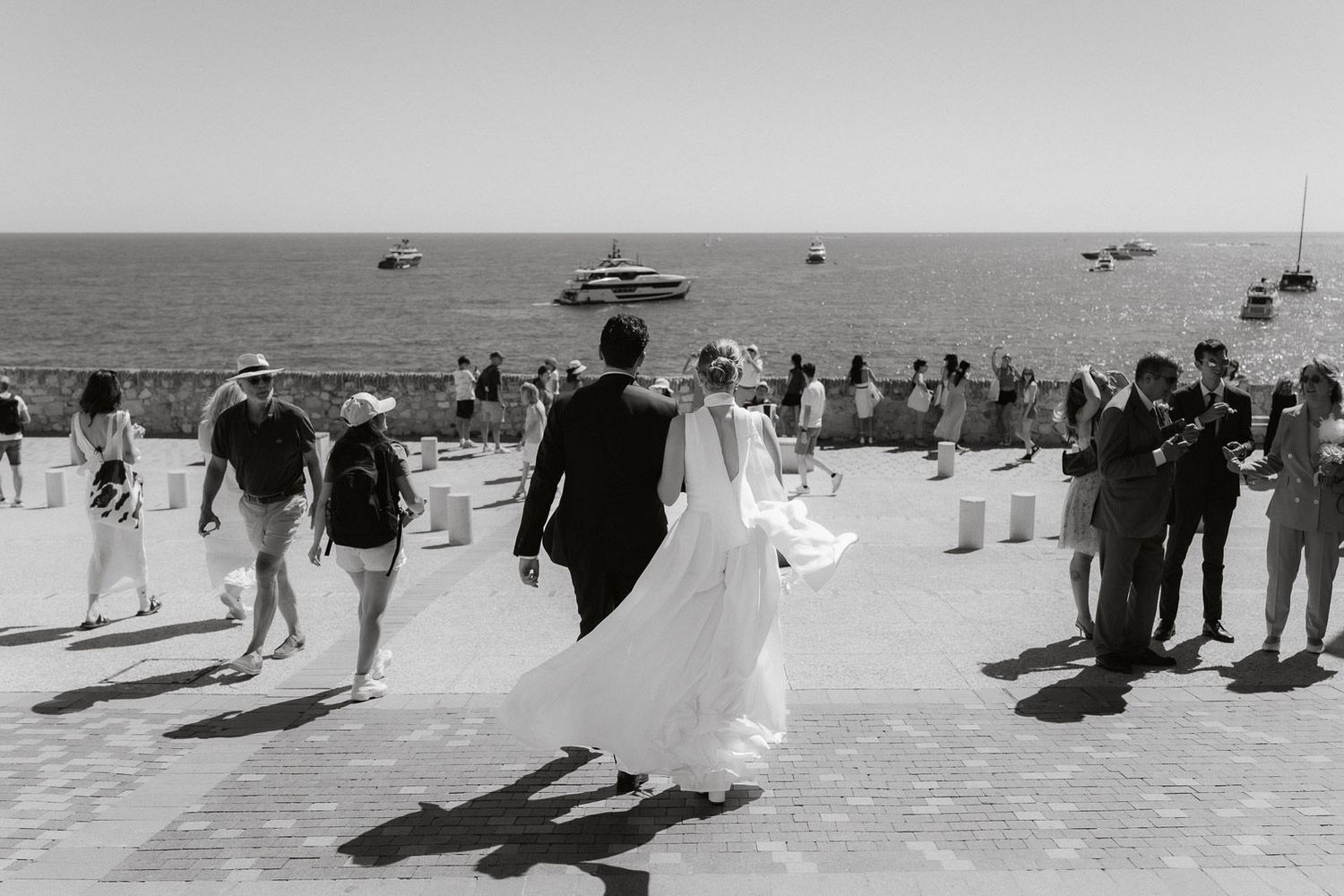 A seaside wedding ceremony with guests gathered on a beach boardwalk as boats float in the distance.