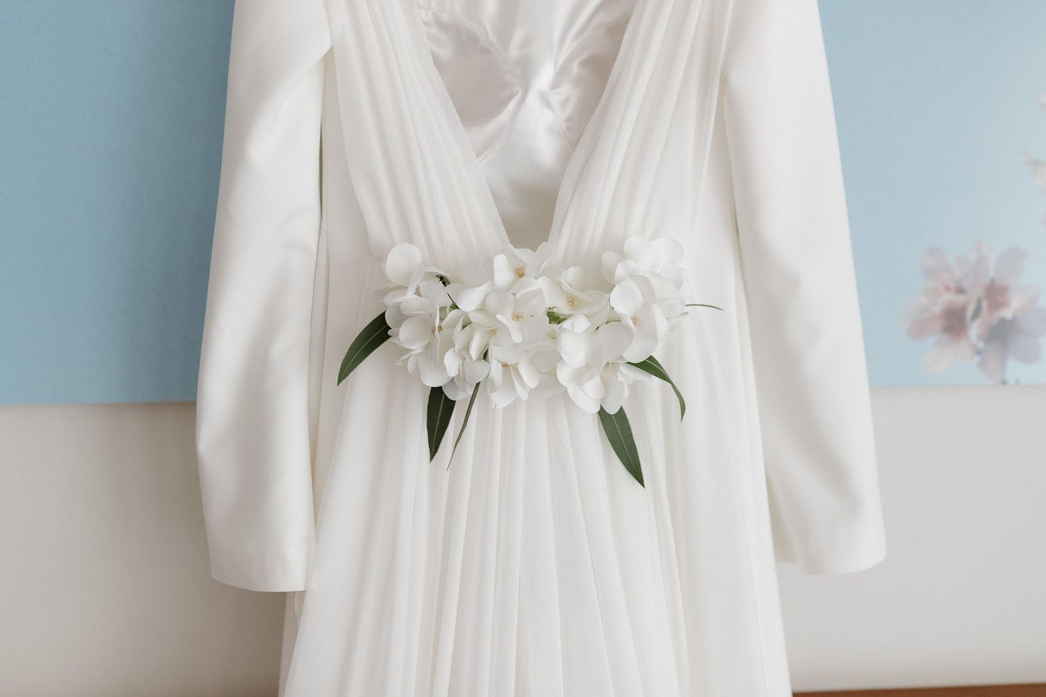A white flowing wedding dress with long sleeves hangs on display, adorned with delicate white lilies at the waist.