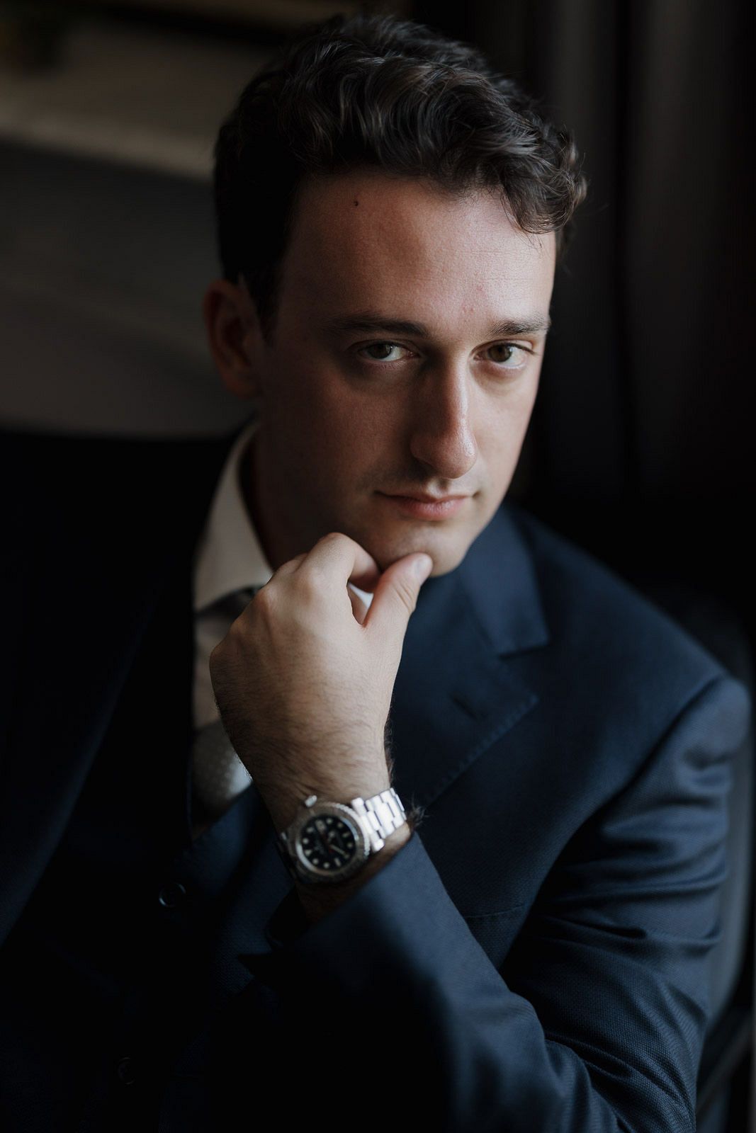A person in a navy suit and white dress shirt poses thoughtfully in moody lighting.