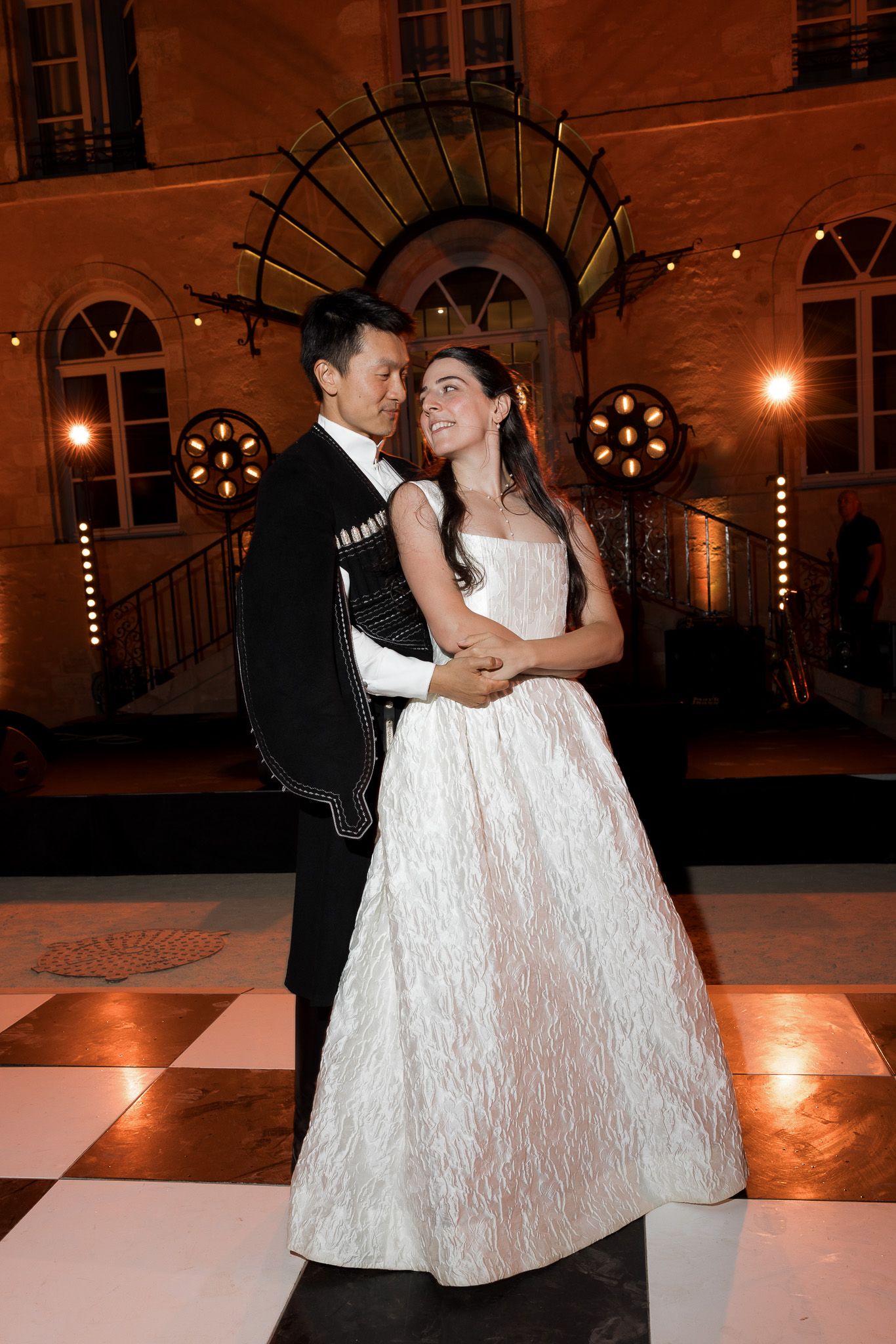 Elegant couple sharing first dance outdoors at night, bride in white gown and groom in black tuxedo on checkered floor.