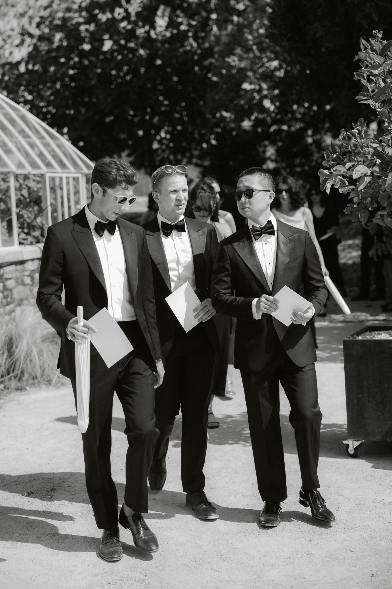 Three groomsmen in tuxedos with bow ties walk together holding ceremony programs at an outdoor wedding.