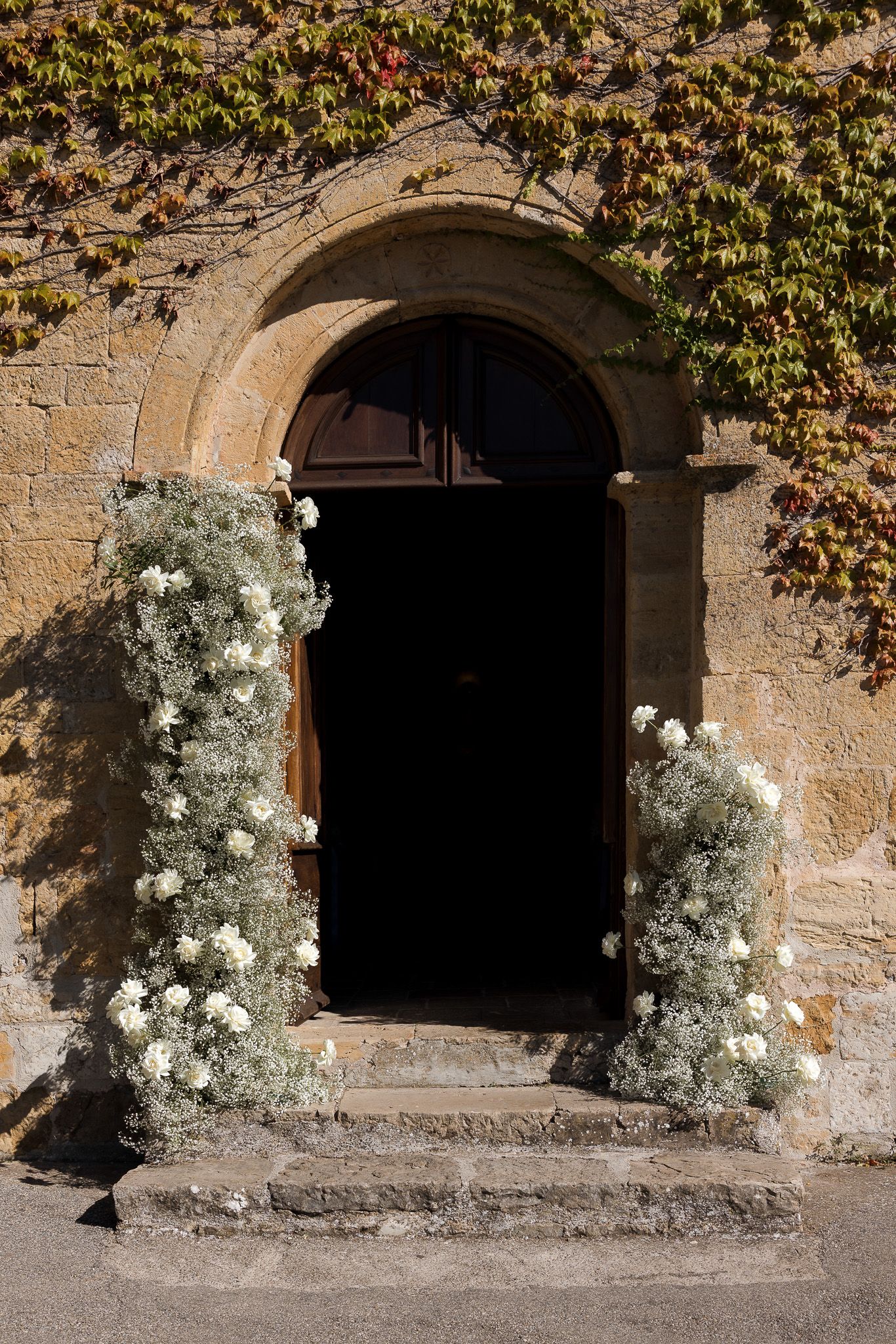 Stone arched doorway adorned with white floral arrangements and ivy-covered walls in warm autumn sunlight.