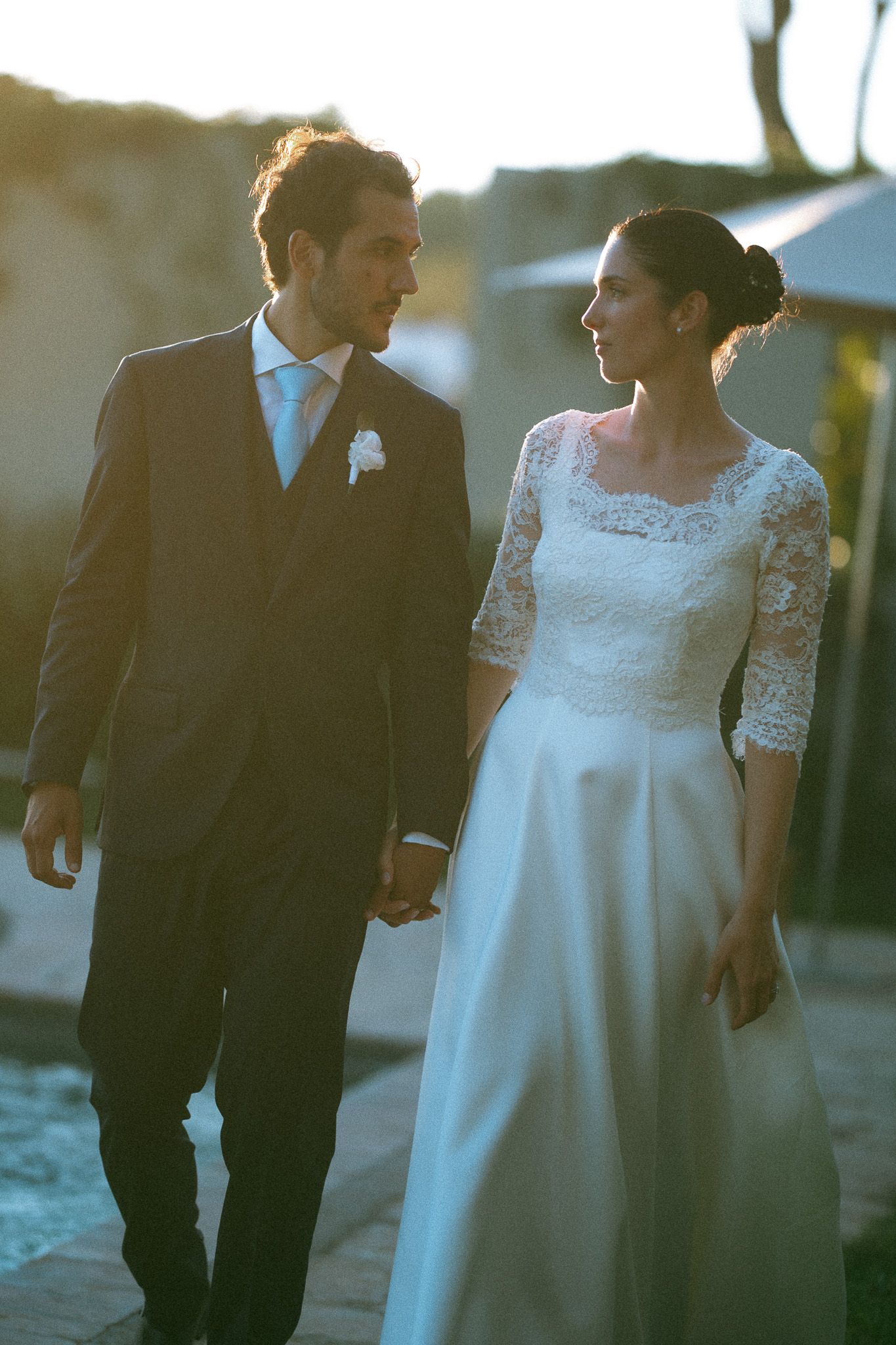 Newlyweds holding hands and walking together at sunset, bride in lace gown and groom in dark suit with boutonniere.