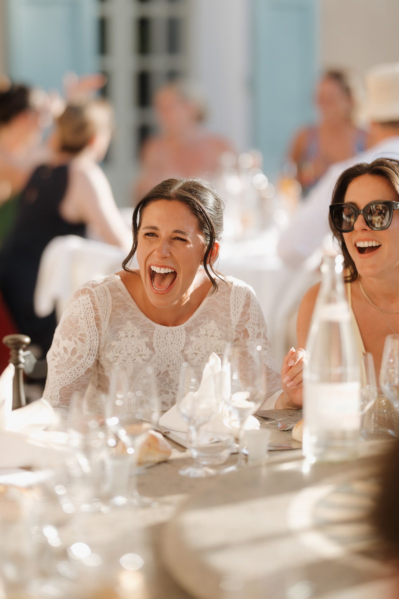Two women laughing joyfully at a wedding reception table, one in white lace dress, elegant dining setting.