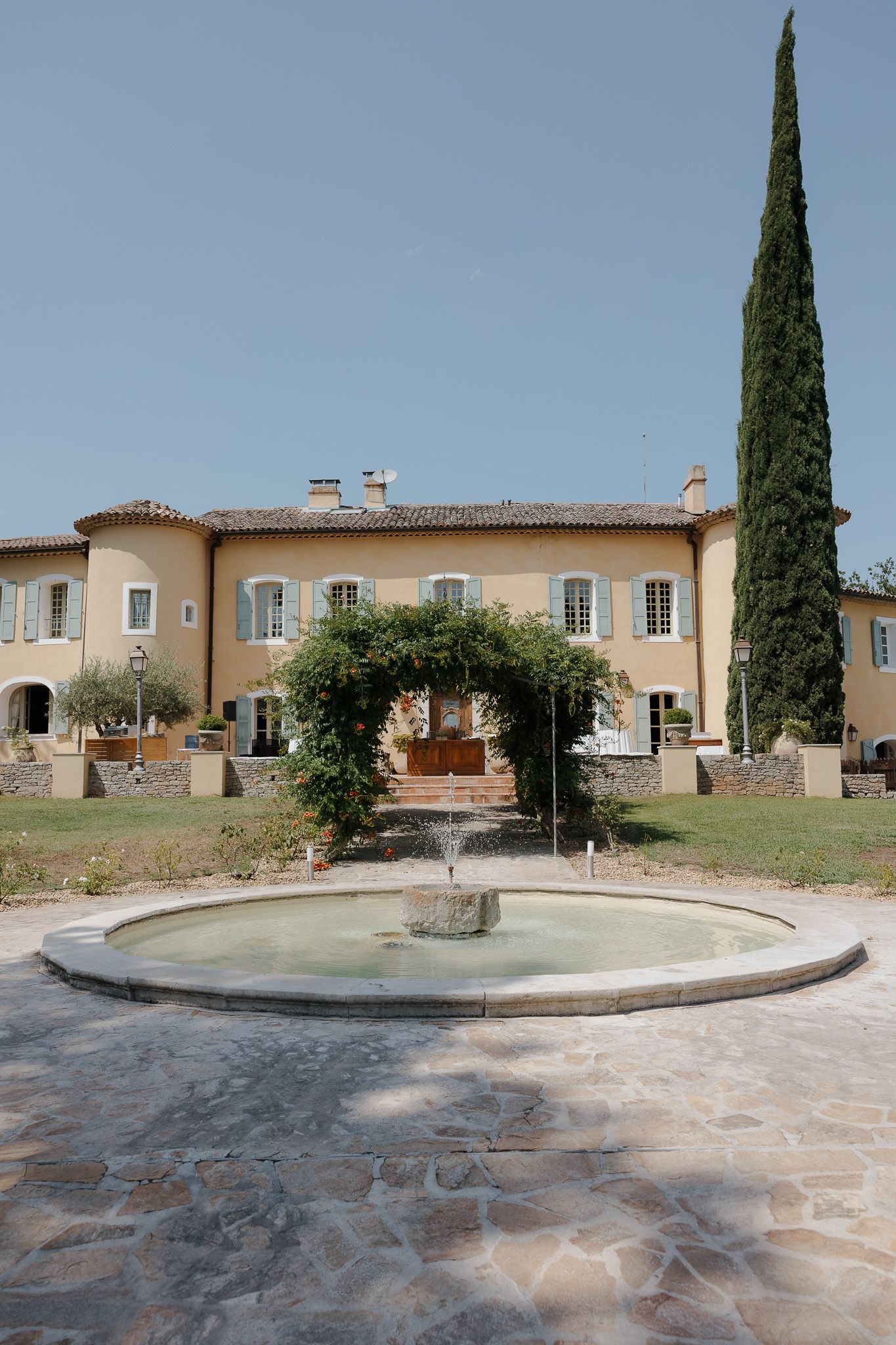 Elegant French Provençal villa with yellow facade, blue shutters, circular fountain, and tall cypress tree under clear blue sky.