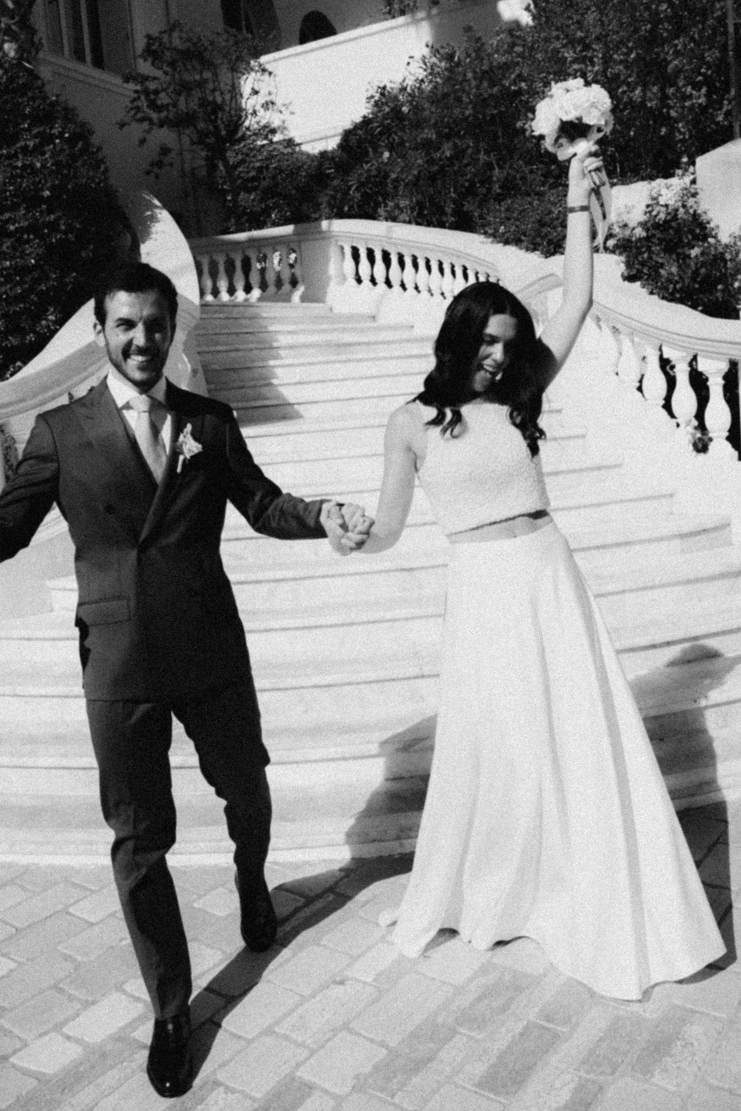 Black and white photo of a joyful bride and groom holding hands on elegant stone steps after their wedding ceremony.