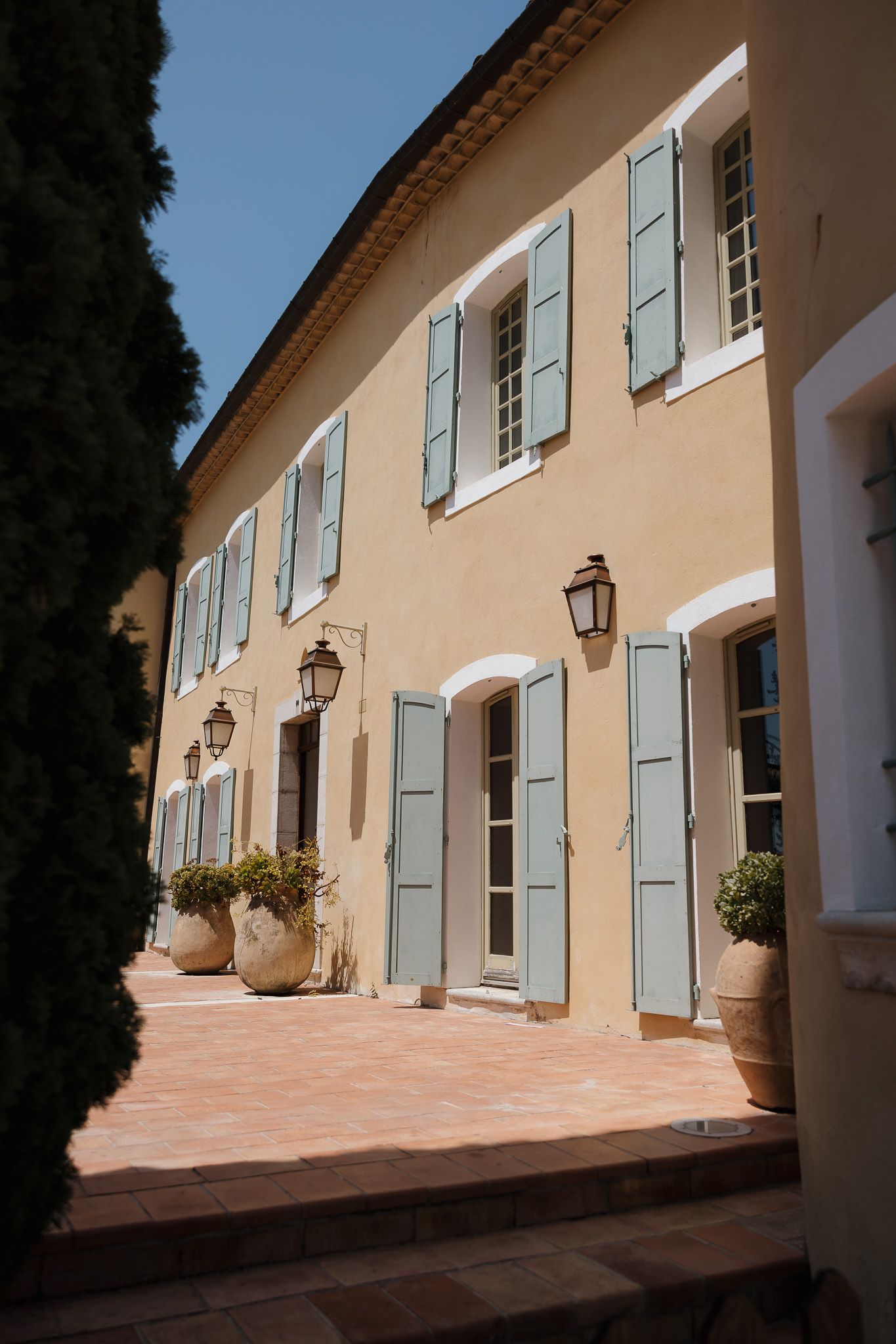 Provençal yellow building with blue shutters, terracotta patio, clay pots, and wall lanterns under a clear blue sky.