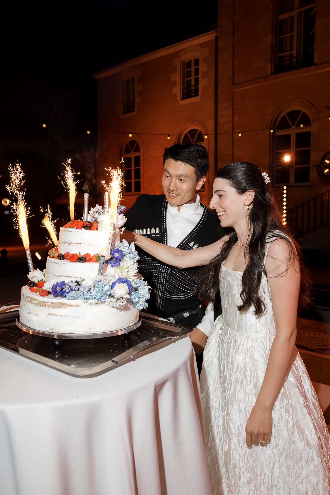 Bride and groom cutting a tiered wedding cake with sparklers outdoors at night near a lit building.