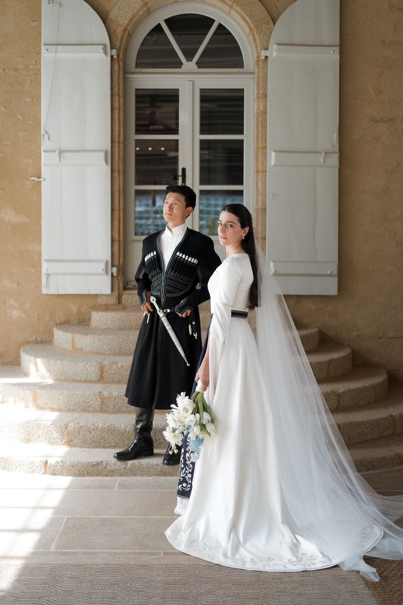 Bride in white gown holding flowers stands with groom in traditional black attire on stone steps of elegant venue.