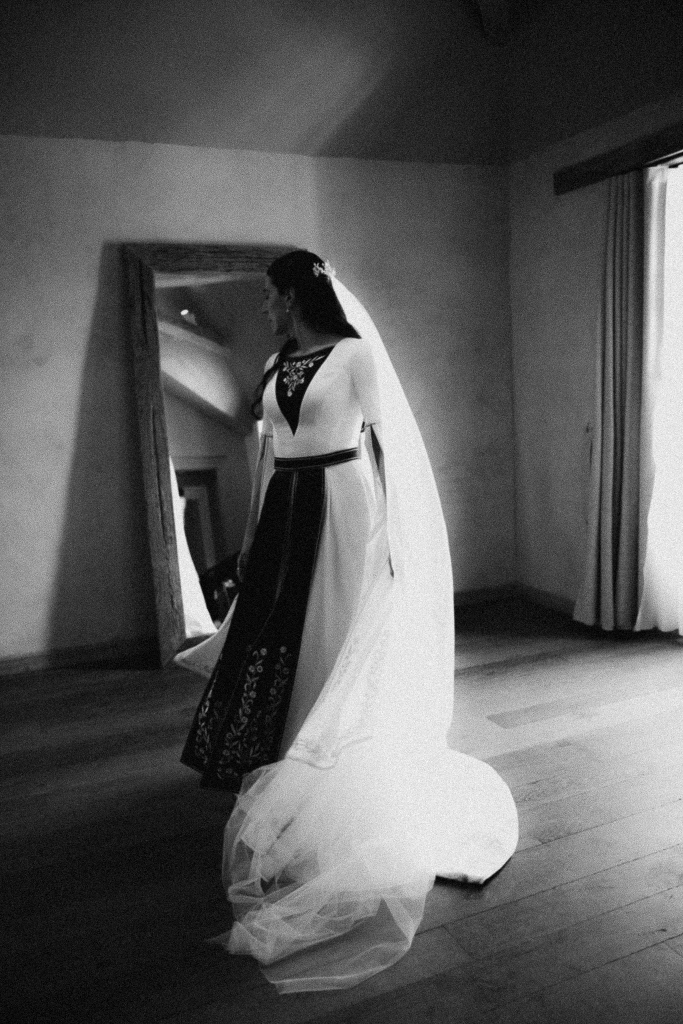 Black and white photo of a bride in a wedding gown with veil standing before a mirror in a softly lit room.