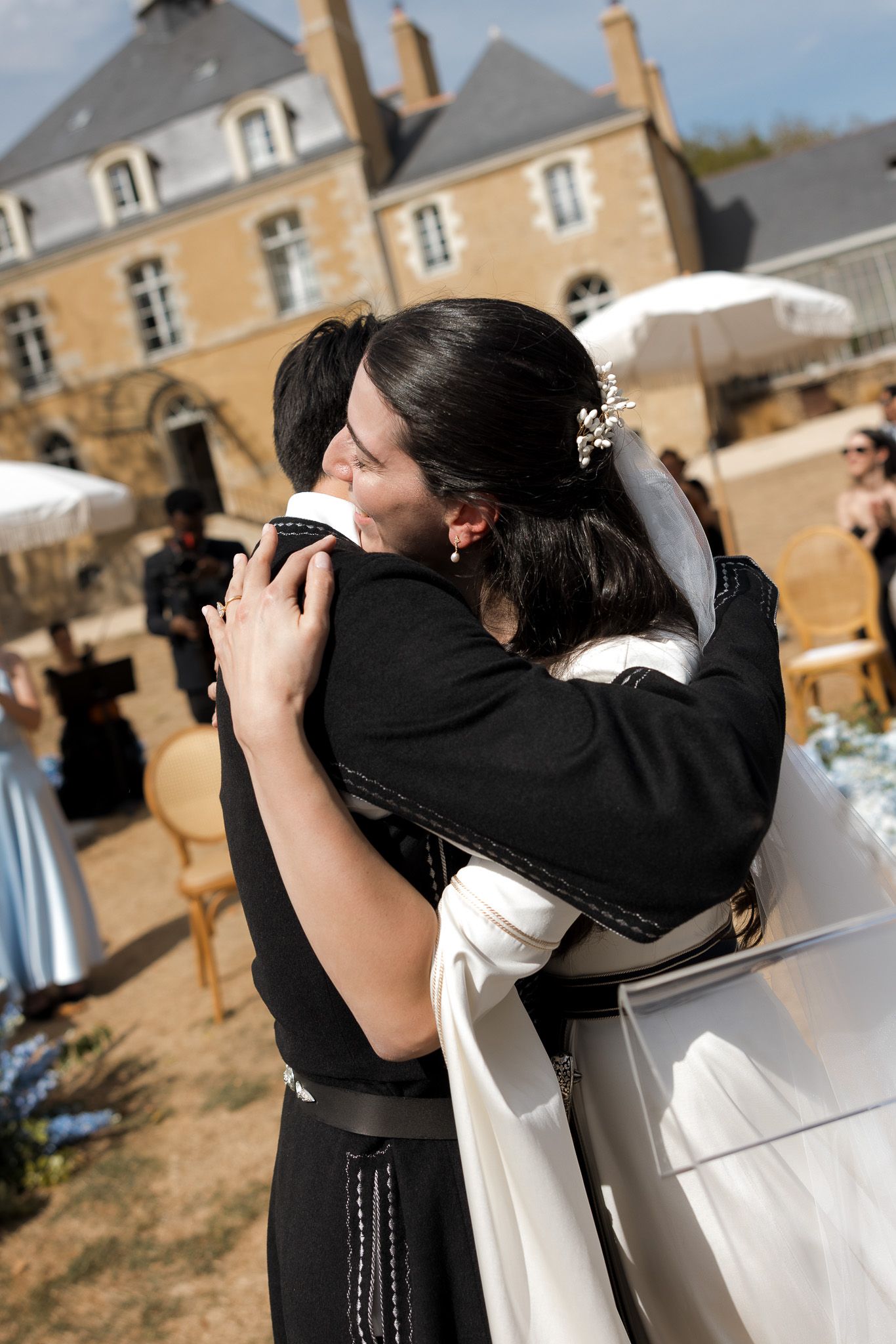 Bride and groom share a romantic embrace outdoors at a wedding ceremony in front of a French chateau.