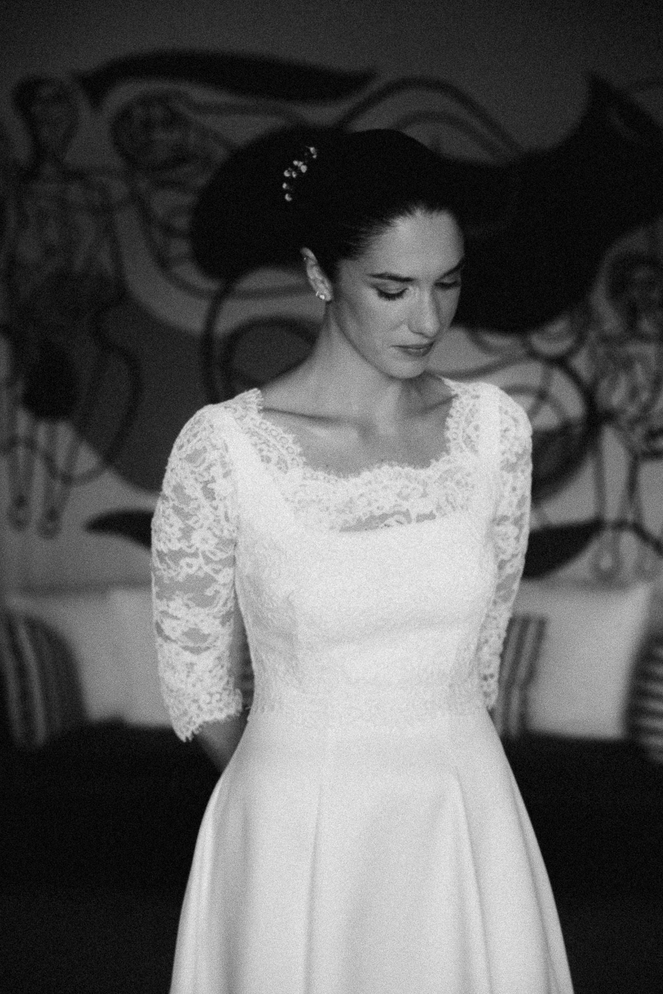 Black and white photo of a bride in a lace sleeve wedding gown with hair accessory, looking down elegantly.