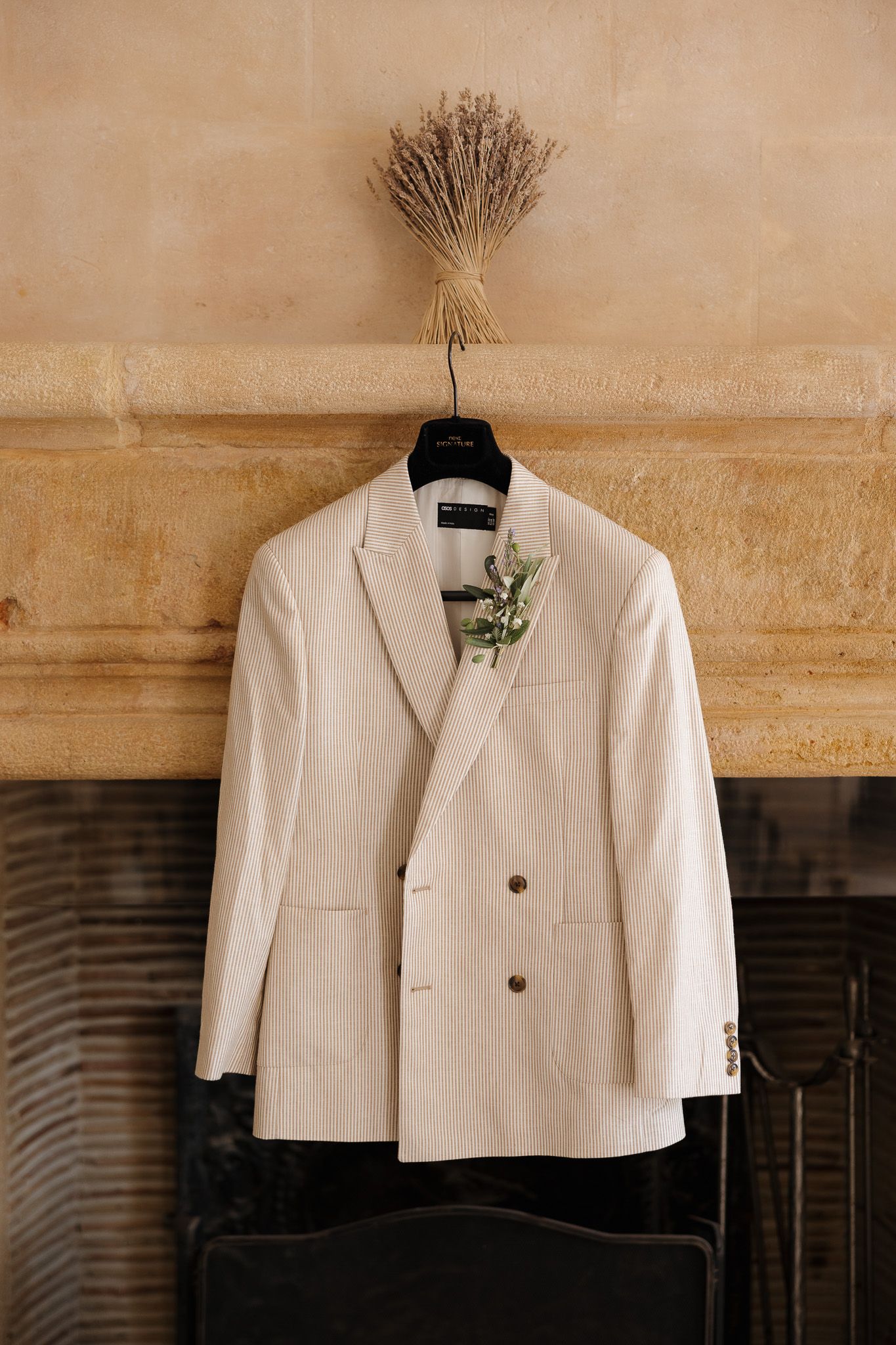 Ivory double-breasted wedding suit jacket with boutonniere hanging on fireplace mantel with dried flowers.
