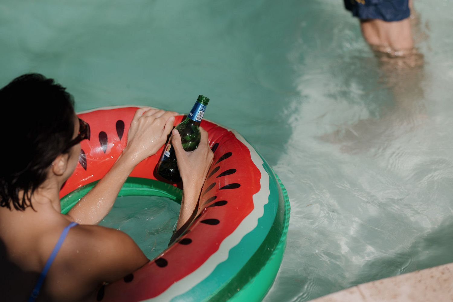 Person floating in pool with inflatable watermelon ring tube on a summer day.