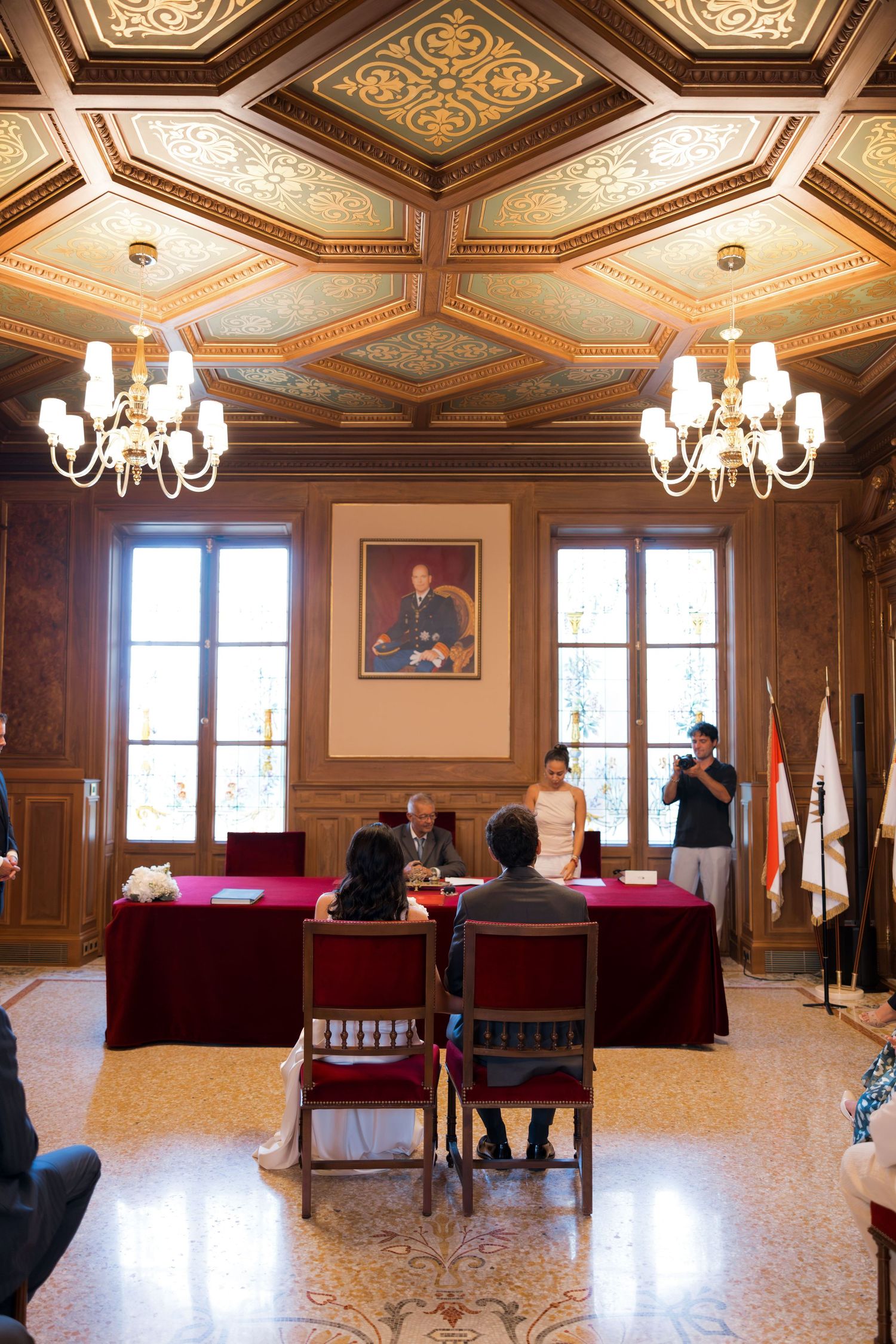 Elegant wedding ceremony in ornate wood-paneled hall with chandelier, couple at red-draped table beneath portrait.