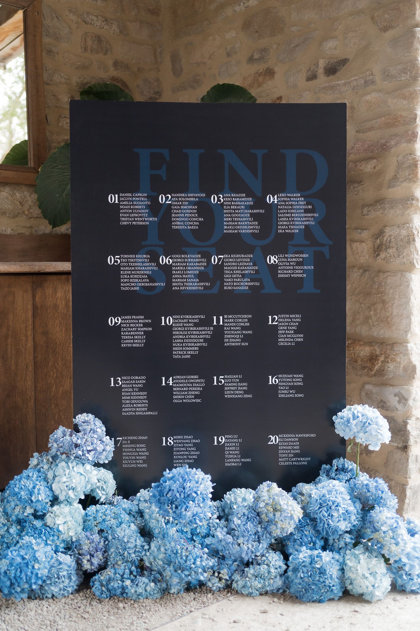 Elegant wedding seating chart on dark board with blue hydrangeas, reading Find Your Seat, displayed against stone wall.