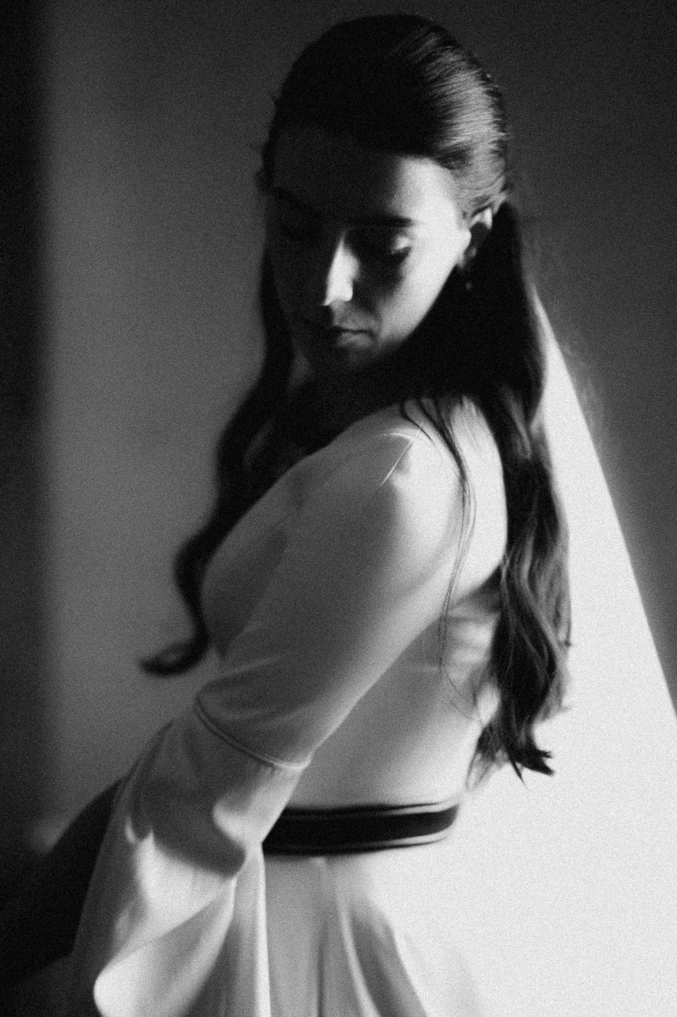 Black and white portrait of a young woman in a white belted dress with long wavy hair, looking down in dramatic light.