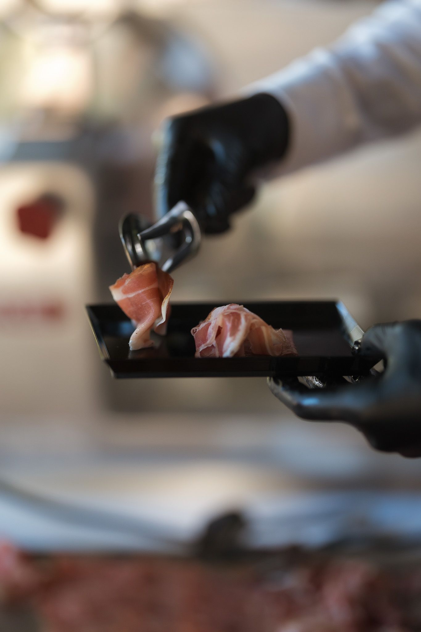 Chef in black gloves placing prosciutto slices on a black serving tray in a professional kitchen.