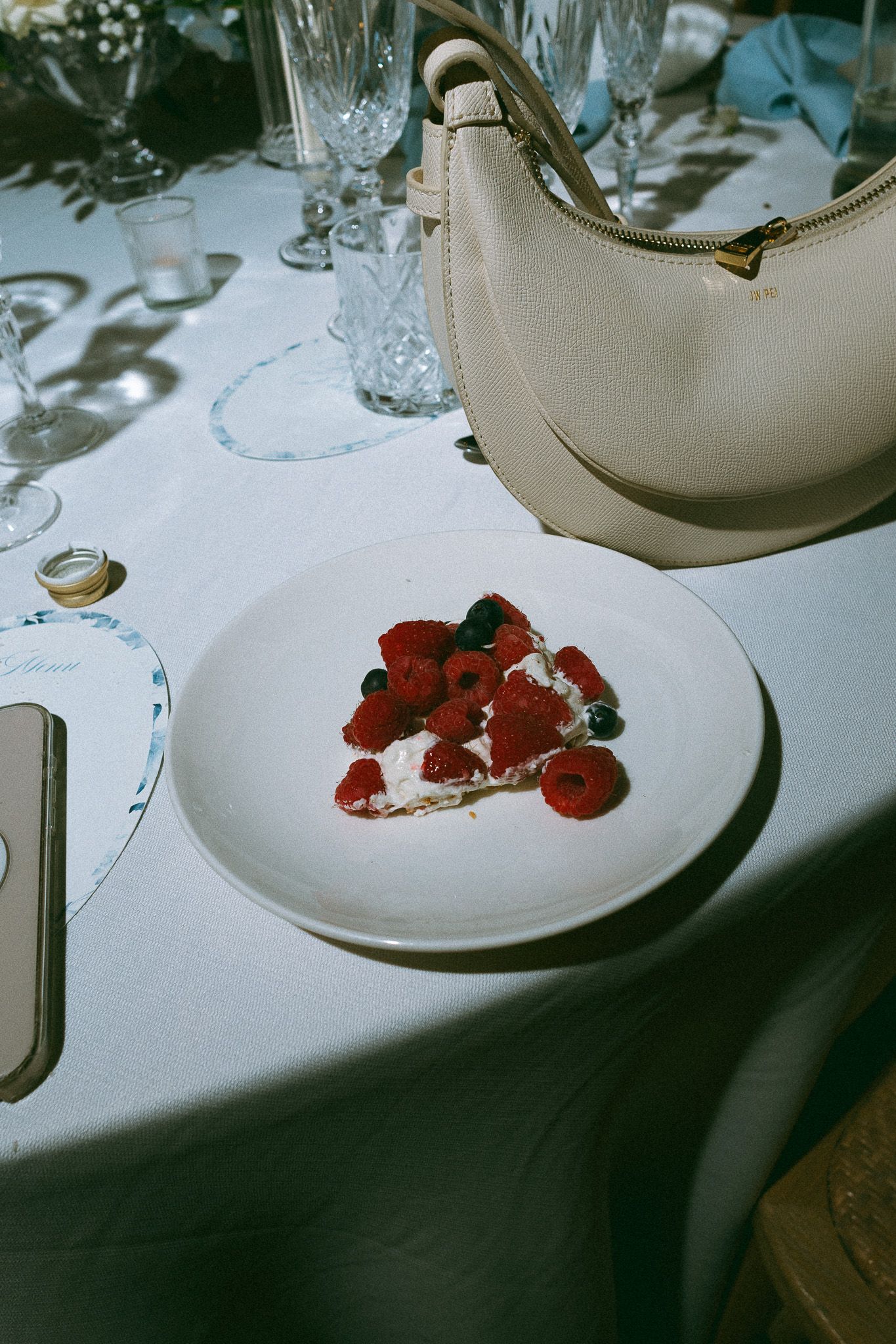 A dessert plate with fresh raspberries and blueberries on cream, beside a beige handbag on a white tablecloth.