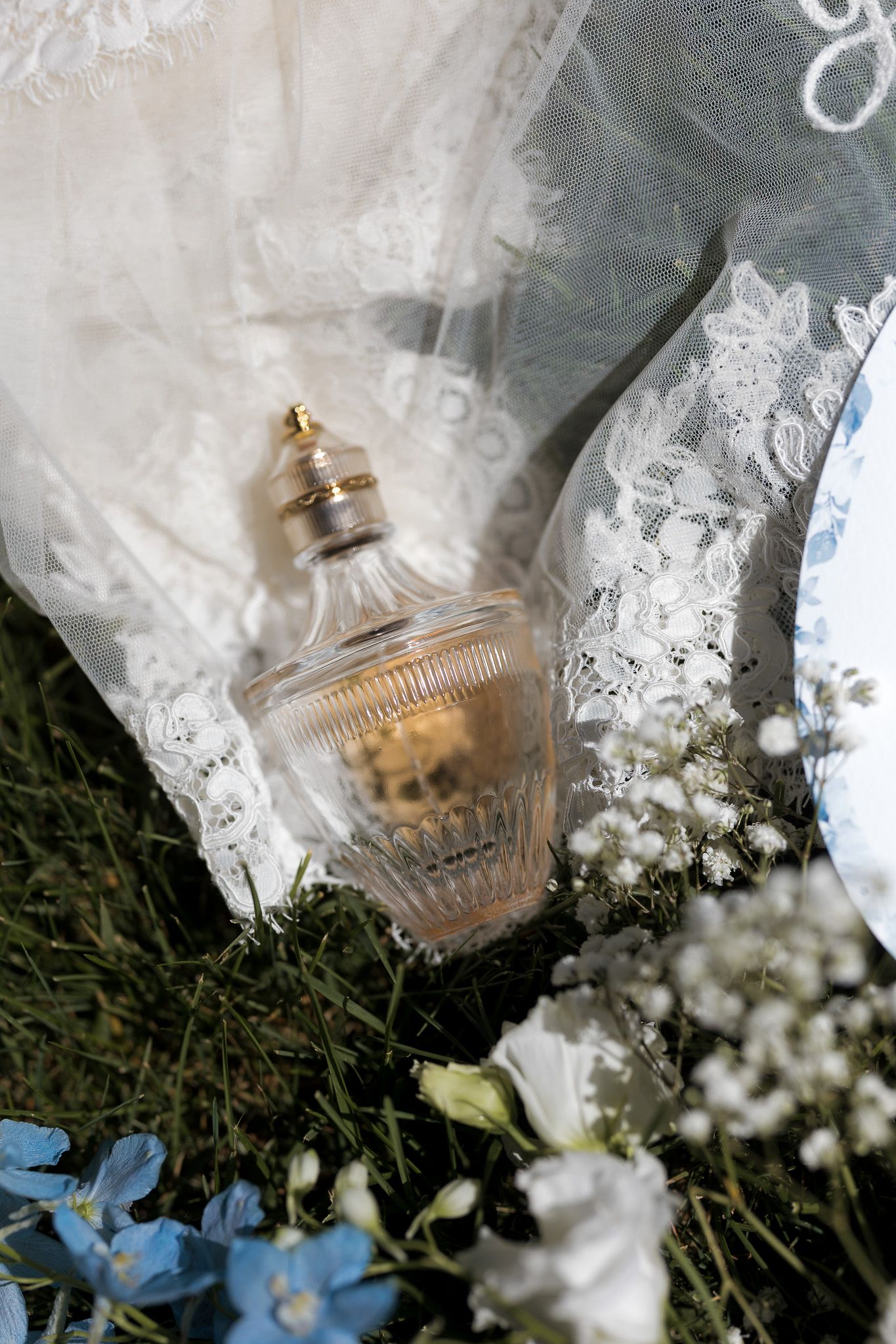 Elegant perfume bottle resting on lace bridal veil surrounded by blue and white wildflowers on green grass.
