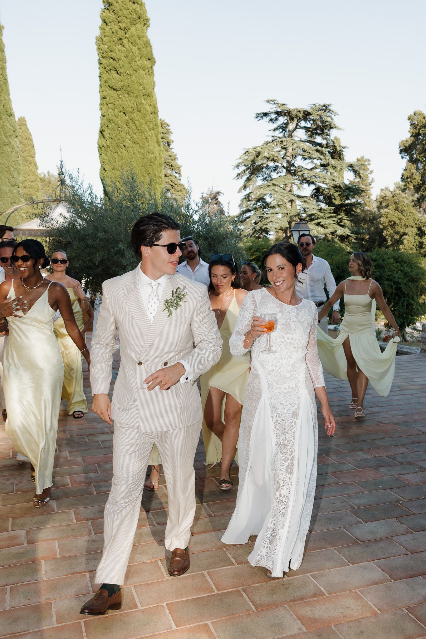 A bride and groom walk joyfully with wedding guests outdoors, surrounded by cypress trees at a sunny celebration.