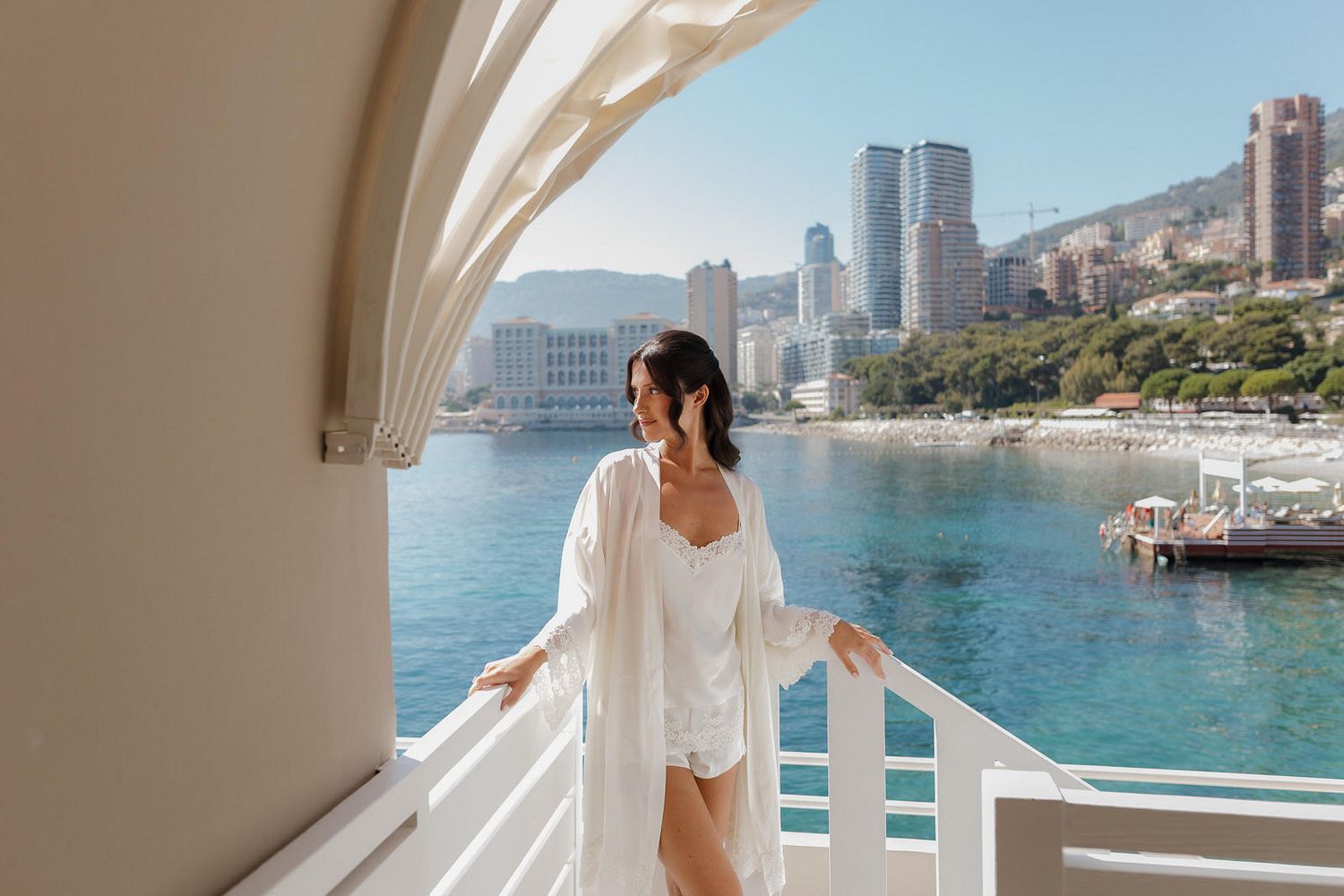 Woman in white dress walking along Mediterranean coastal promenade with luxury buildings and blue sea views.