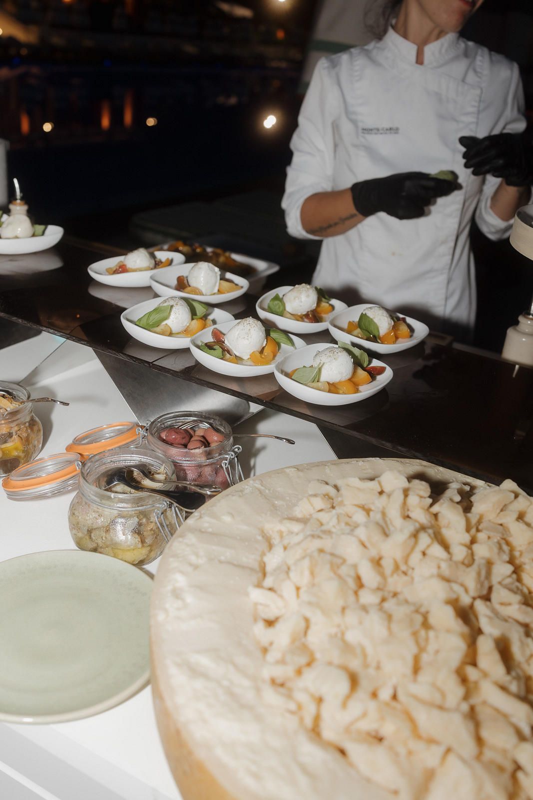 Chef in white uniform plating elegant dishes at a catering event with appetizers and large cheese wheel in foreground.