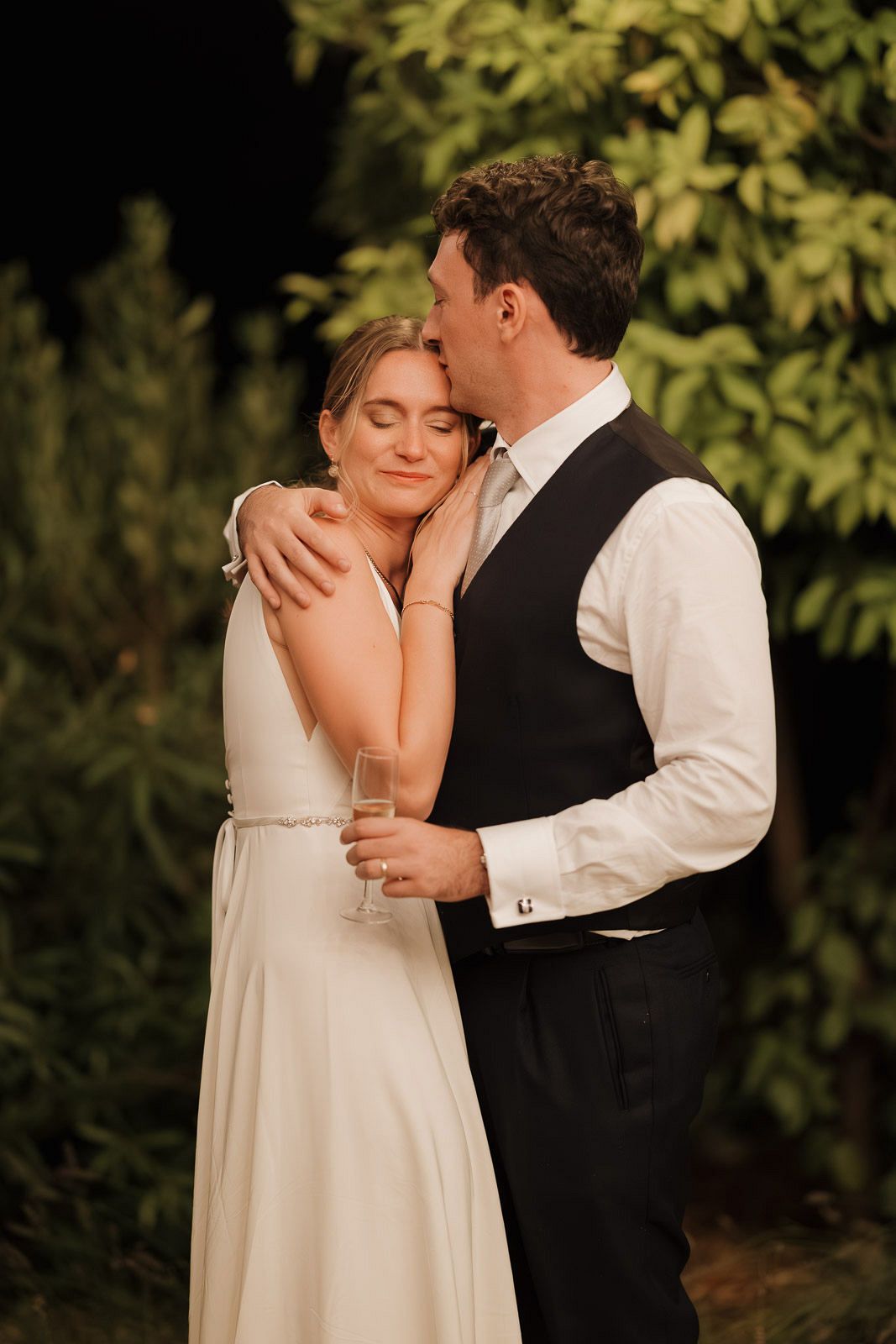 A romantic wedding couple embraces in a garden setting with lush greenery in the background.