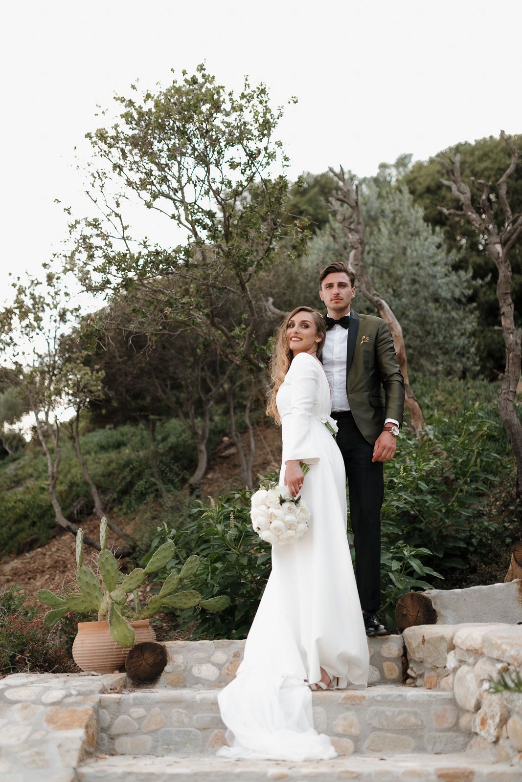 A wedding couple shares romantic moments on a stone terrace surrounded by Mediterranean olive trees.