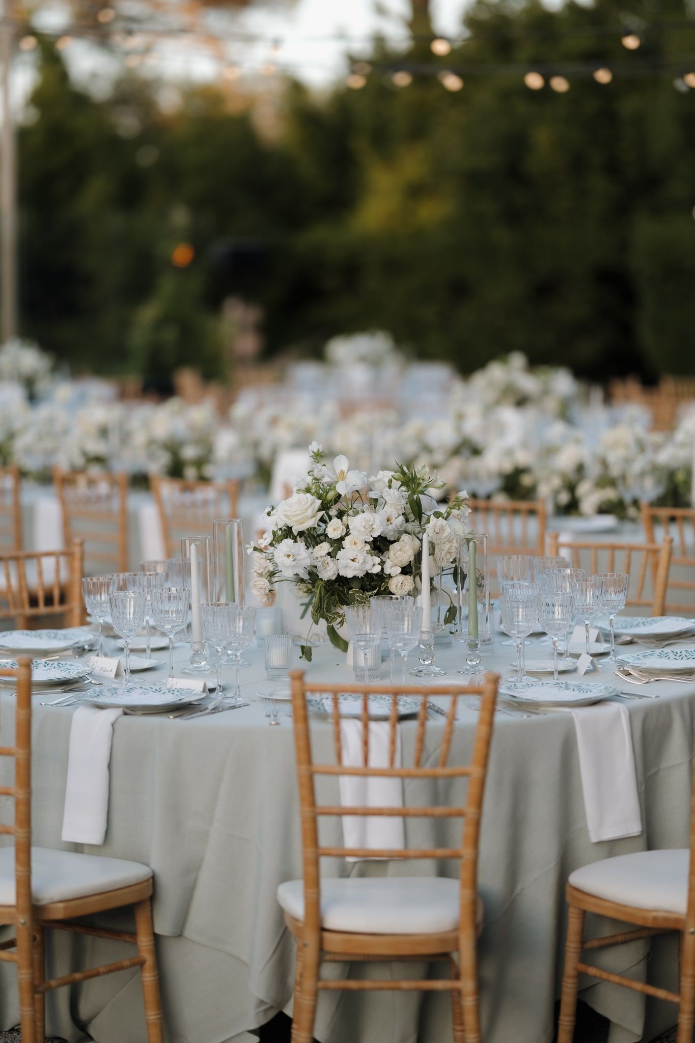 Elegant outdoor wedding reception setup with white tablecloths, wooden chairs and delicate floral centerpieces at dusk.