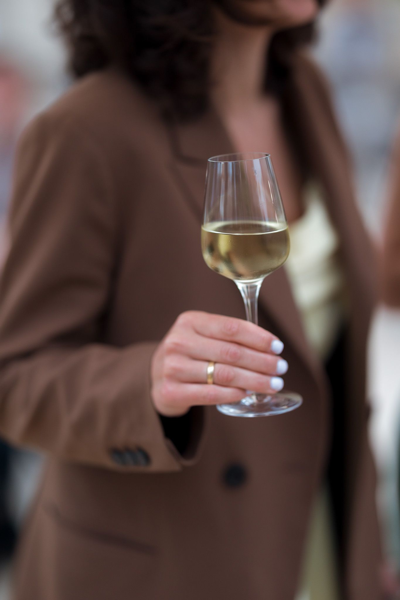 Woman in brown blazer holding a glass of white wine at a social event.