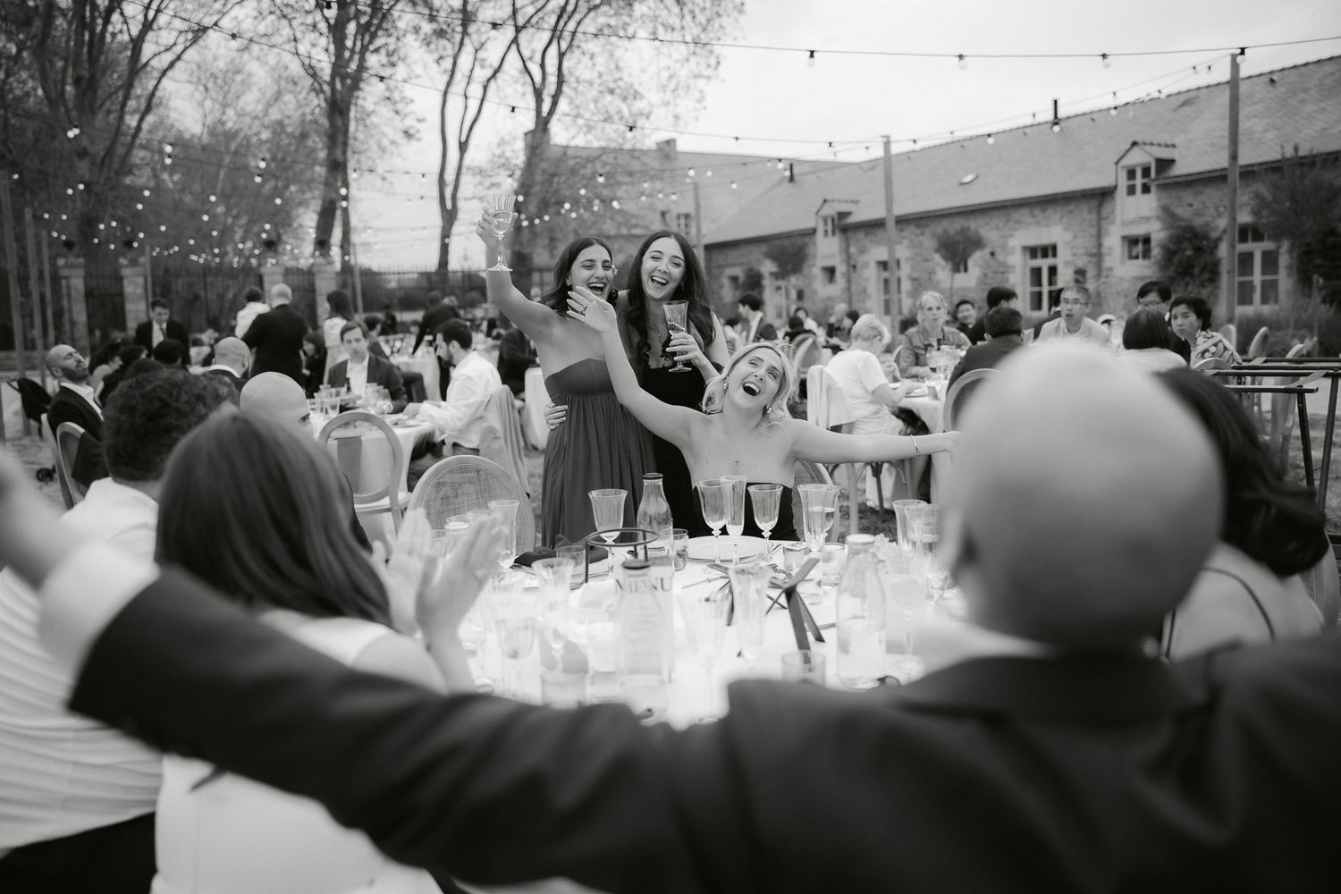 Black and white wedding reception photo of guests celebrating outdoors under string lights at elegant venue.