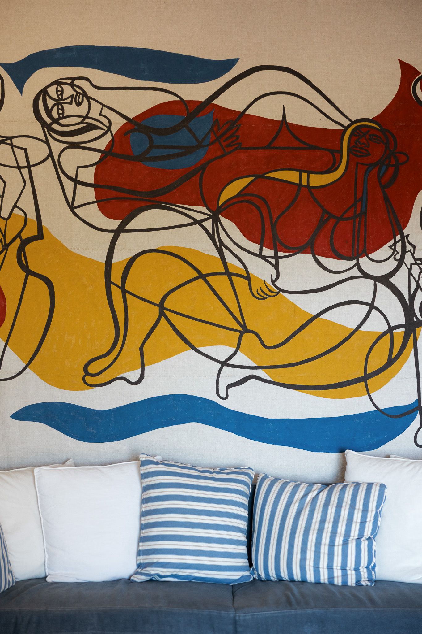 Abstract mural with bold red, yellow, and blue figures above a gray sofa with striped cushions.