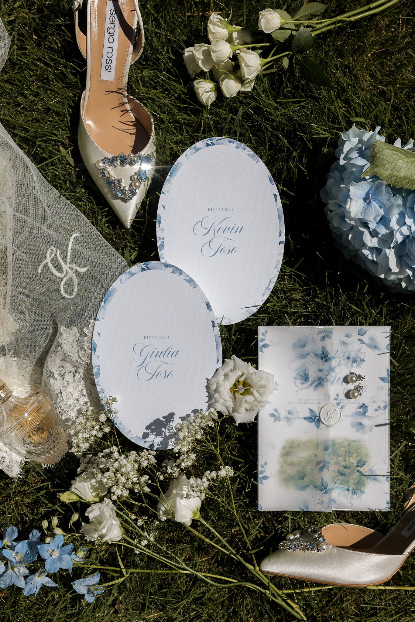 Elegant wedding stationery with blue floral oval cards, bridal shoes, hydrangeas, and delicate white flowers arranged on green grass.