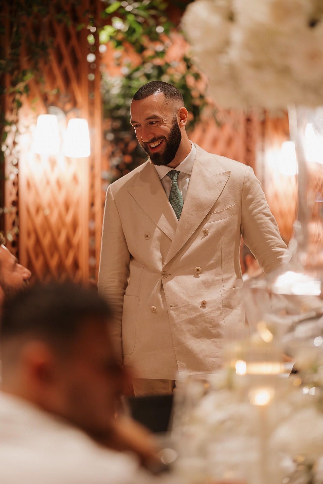 A person in a cream-colored suit jacket smiles warmly at a formal event with soft lighting and decorative elements.
