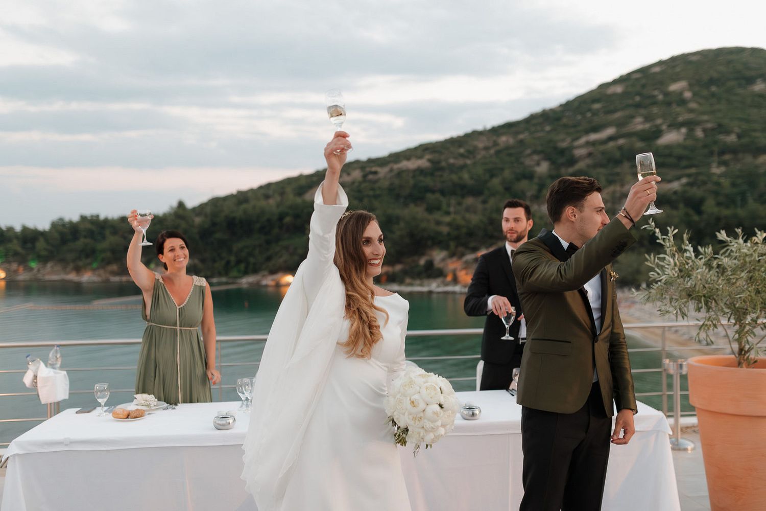 Newlyweds celebrate during sunset by a scenic lakeside setting with mountains in the background.