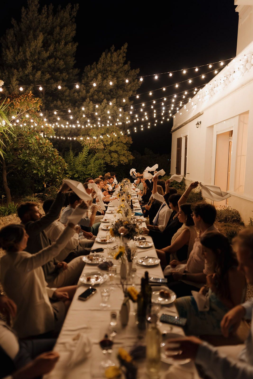 Outdoor dinner party under string lights with guests seated at a long table celebrating at night.