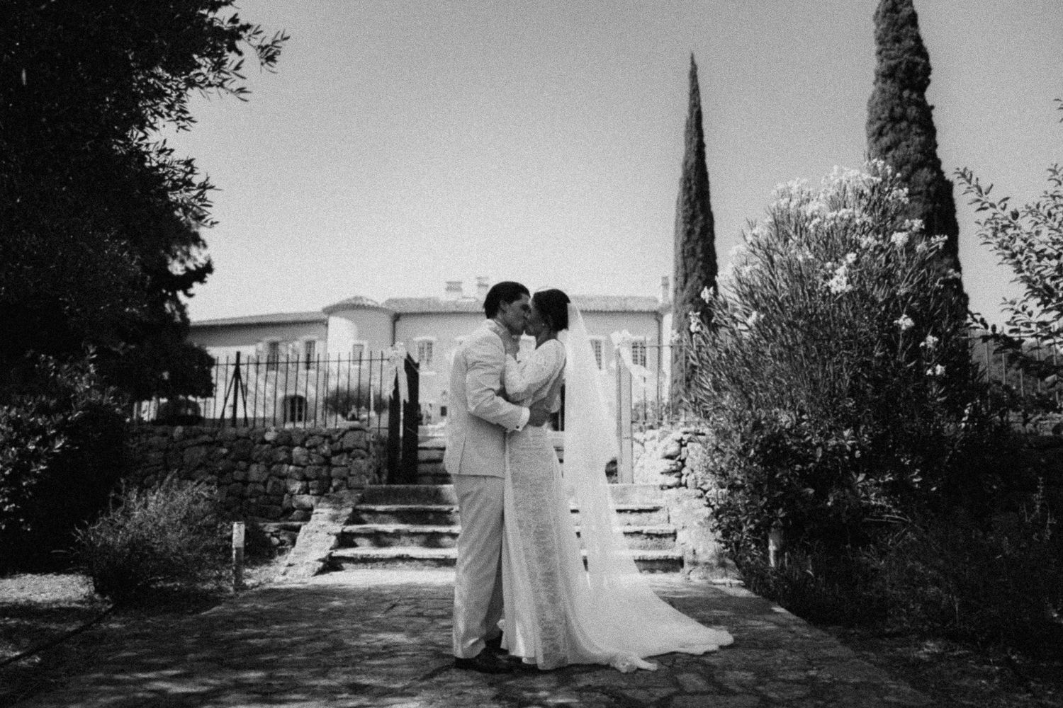 Black and white photo of a bride and groom sharing a kiss outside an elegant villa surrounded by lush gardens.