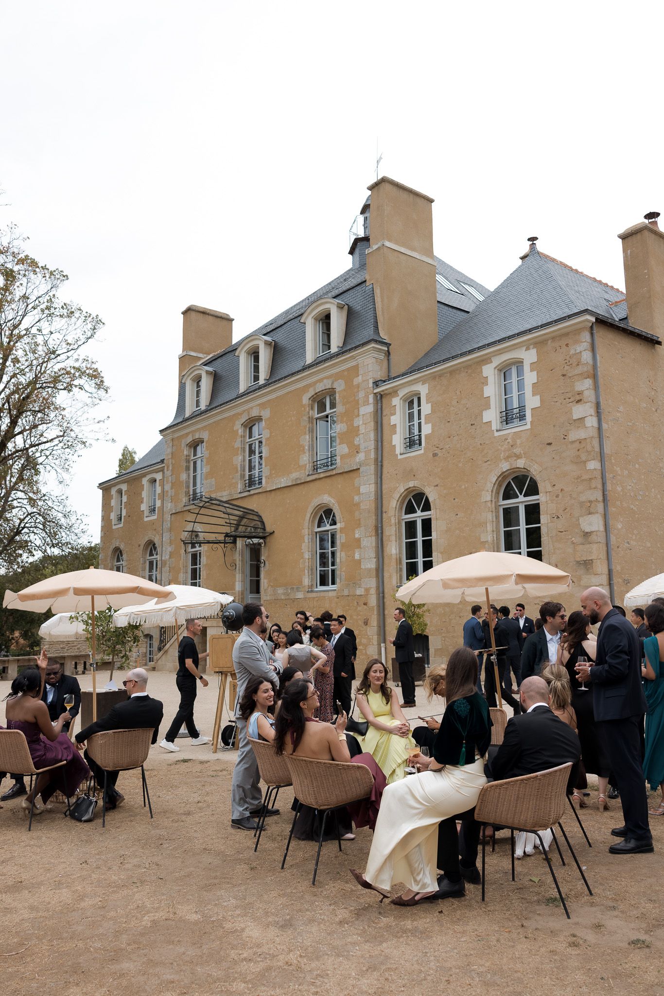 Elegant outdoor wedding reception at a French chateau with guests seated under cream umbrellas on a gravel terrace.