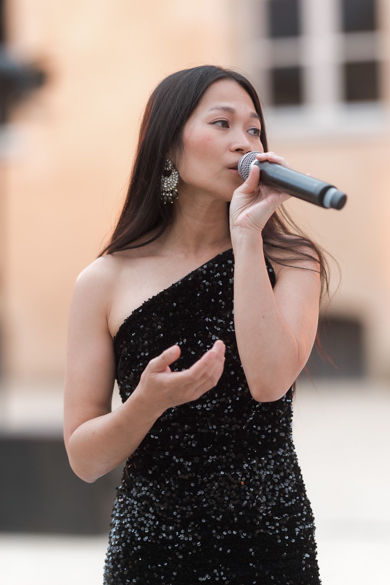 Asian woman in black sequin dress performing with microphone at outdoor event.