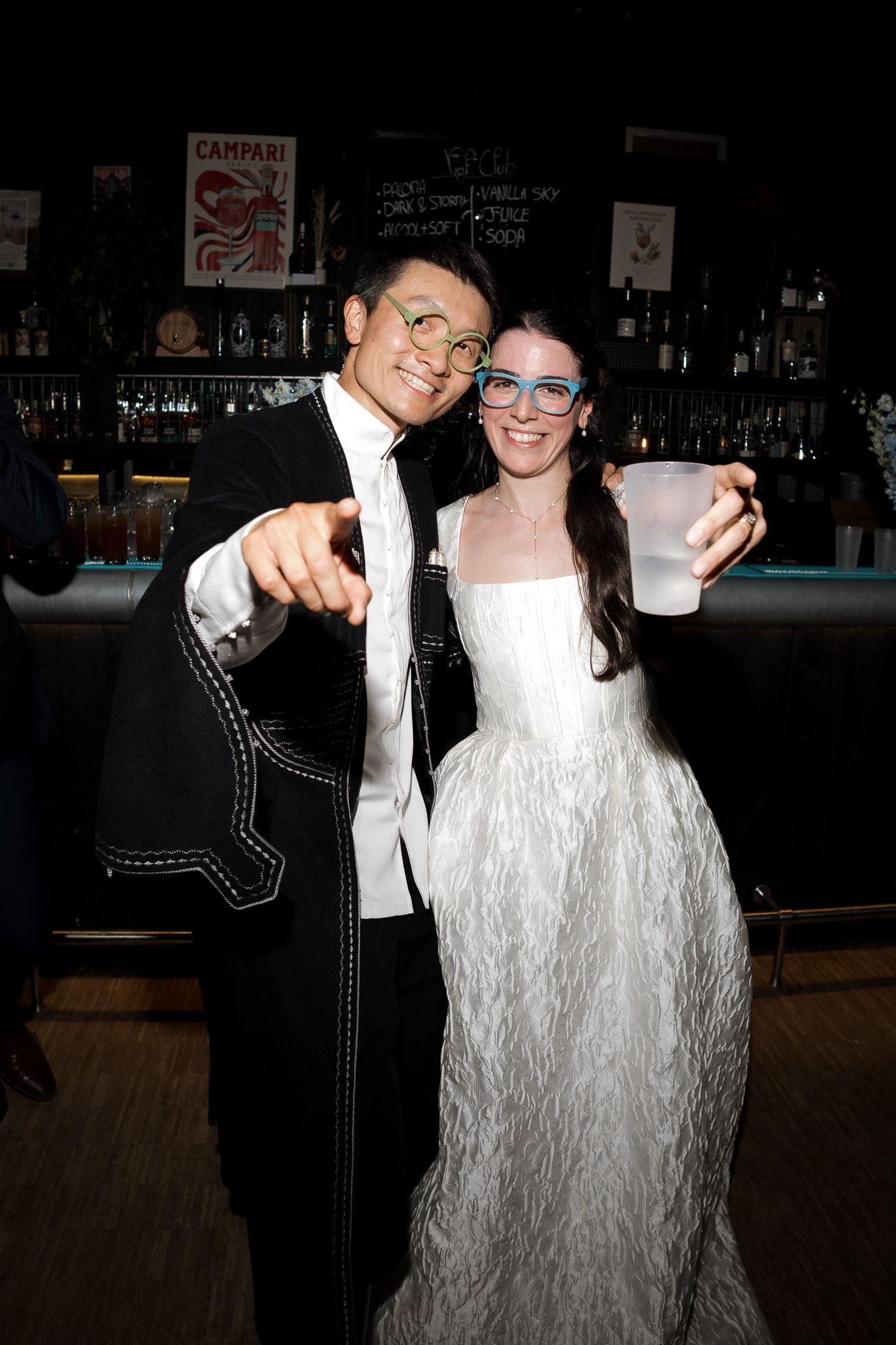 Couple in wedding attire posing at a bar, wearing fun glasses, man pointing at camera, woman holding a plastic cup.