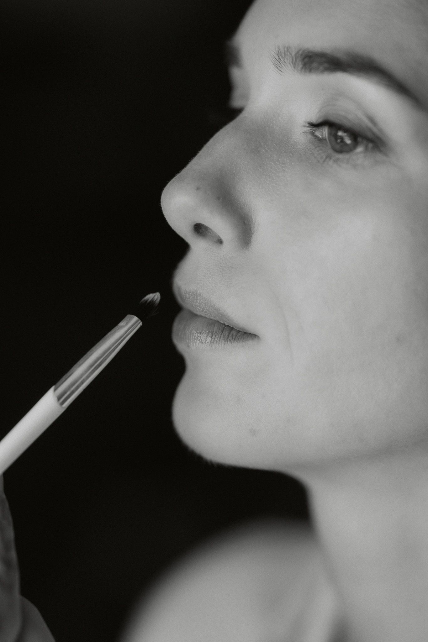Black and white close-up of a woman having lip makeup applied with a brush by a makeup artist.