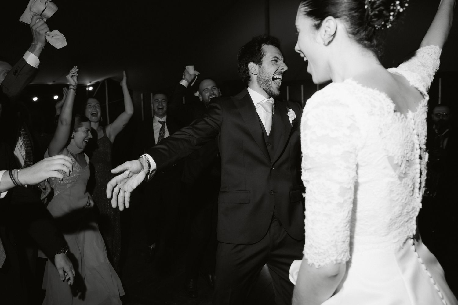 Joyful bride and groom dancing at wedding reception surrounded by celebrating guests in black and white photo.